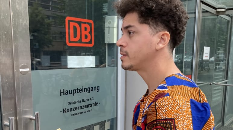 Un jeune homme se tient avec une enveloppe devant une porte vitrée portant l’inscription DB entrée principale siège du groupe Deutsche Bahn AG