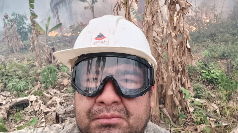 Un homme portant un casque et des lunettes de protection regarde la caméra, tandis que des buissons brûlent et que de la fumée s’élève à l’arrière-plan